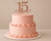 Tarta de 15 años de dos pisos decorada en rosa pastel con rosetones de crema y topper dorado con el número 15, ideal para celebraciones en Vilafranca del Penedès.